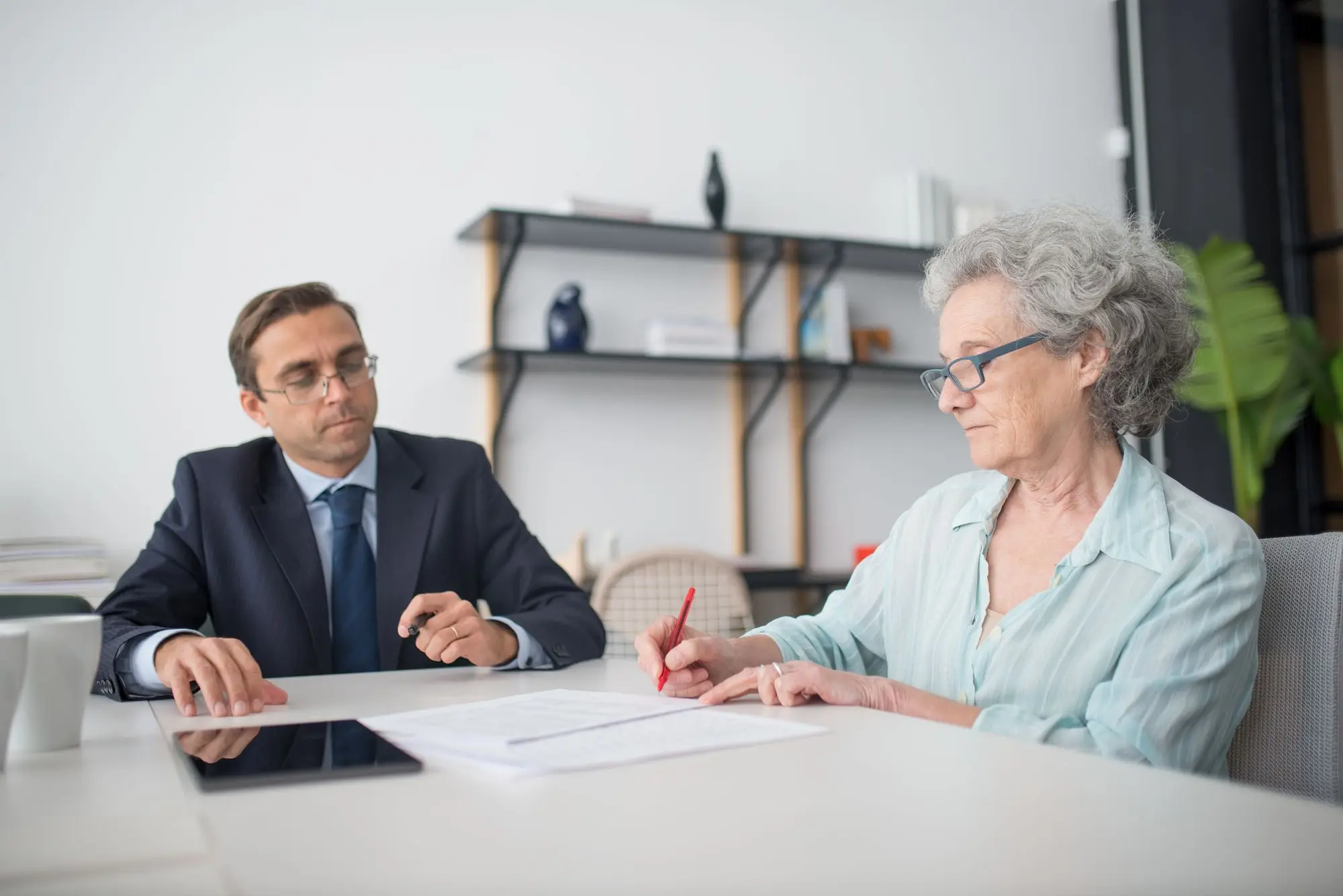 Older business owner signing a loan document at a meeting table while a witness looks on
